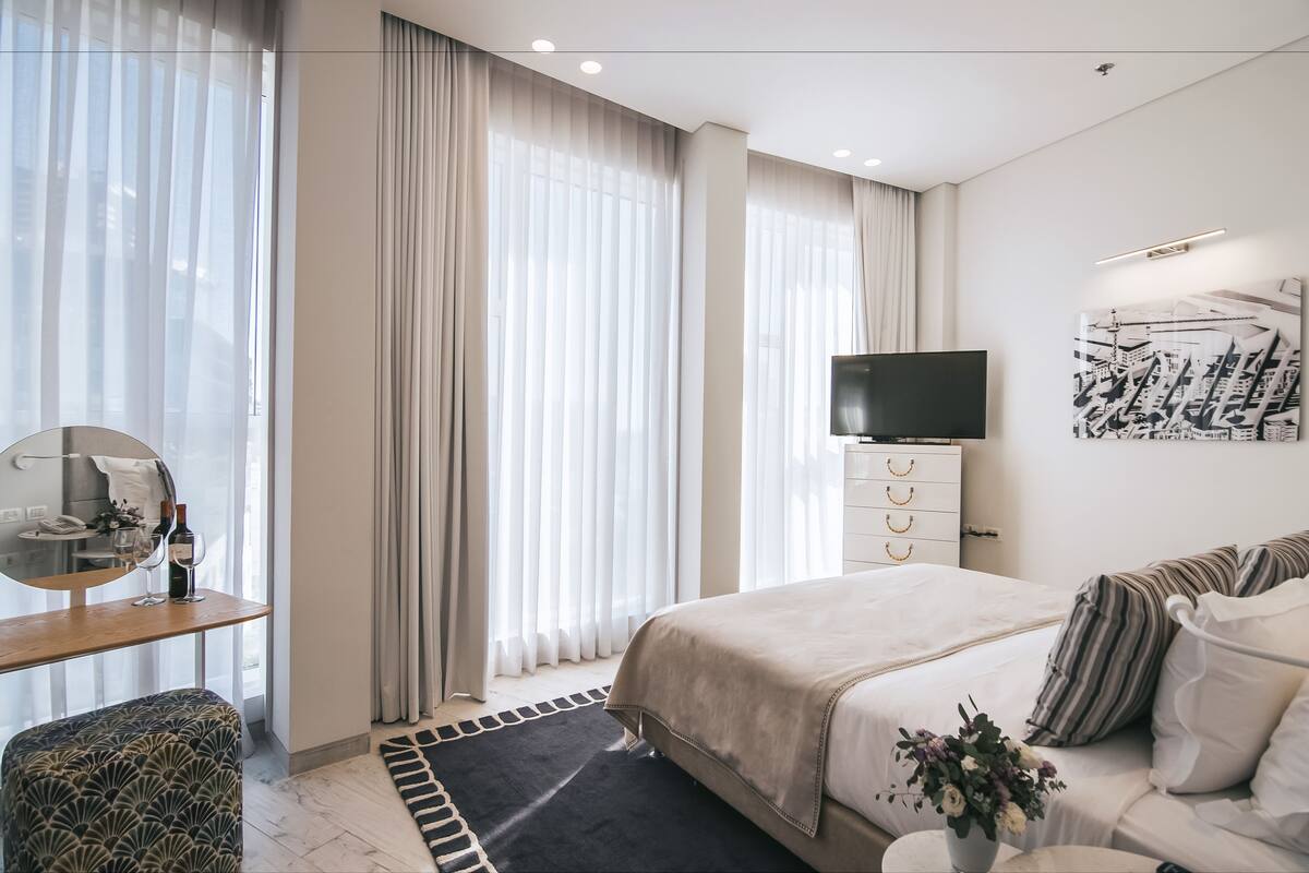 A modern hotel room with a bed, a TV, a desk with a mirror, and a window with curtains.