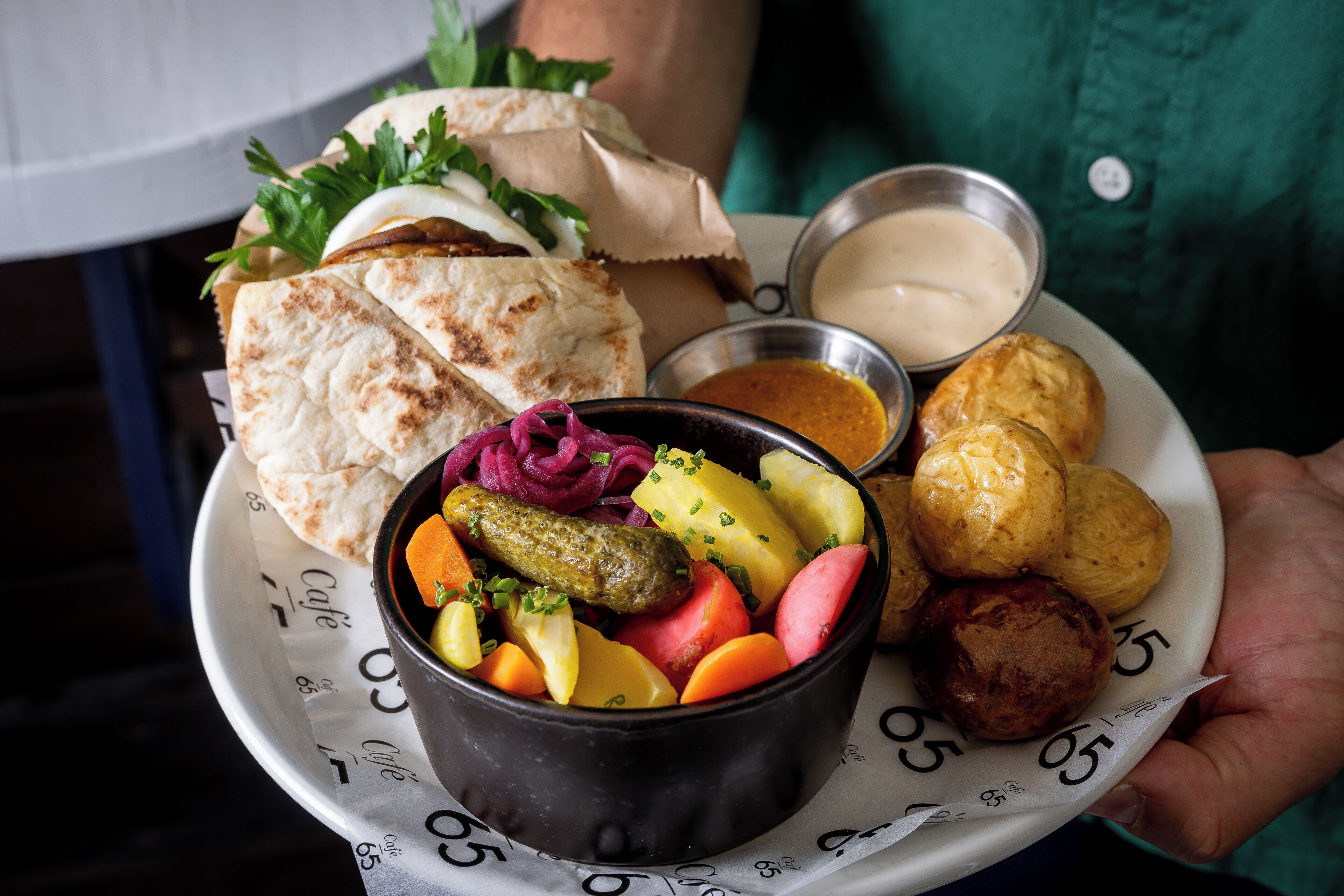 A plate with a variety of foods including bread, pickles, potatoes, and sausages, accompanied by two dipping sauces.