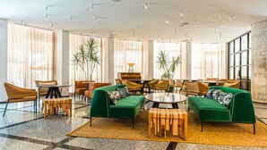 A spacious lounge area with green sofas, wooden tables, and chairs, large windows with sheer curtains, and potted plants. - 65 Hotel, Rothschild Tel Aviv - an Atlas Boutique Hotel (Tel Aviv)