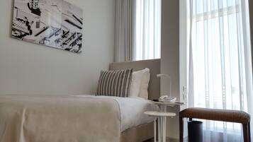 A hotel room with a bed, a bedside table with a phone, a chair, and a large window with curtains.