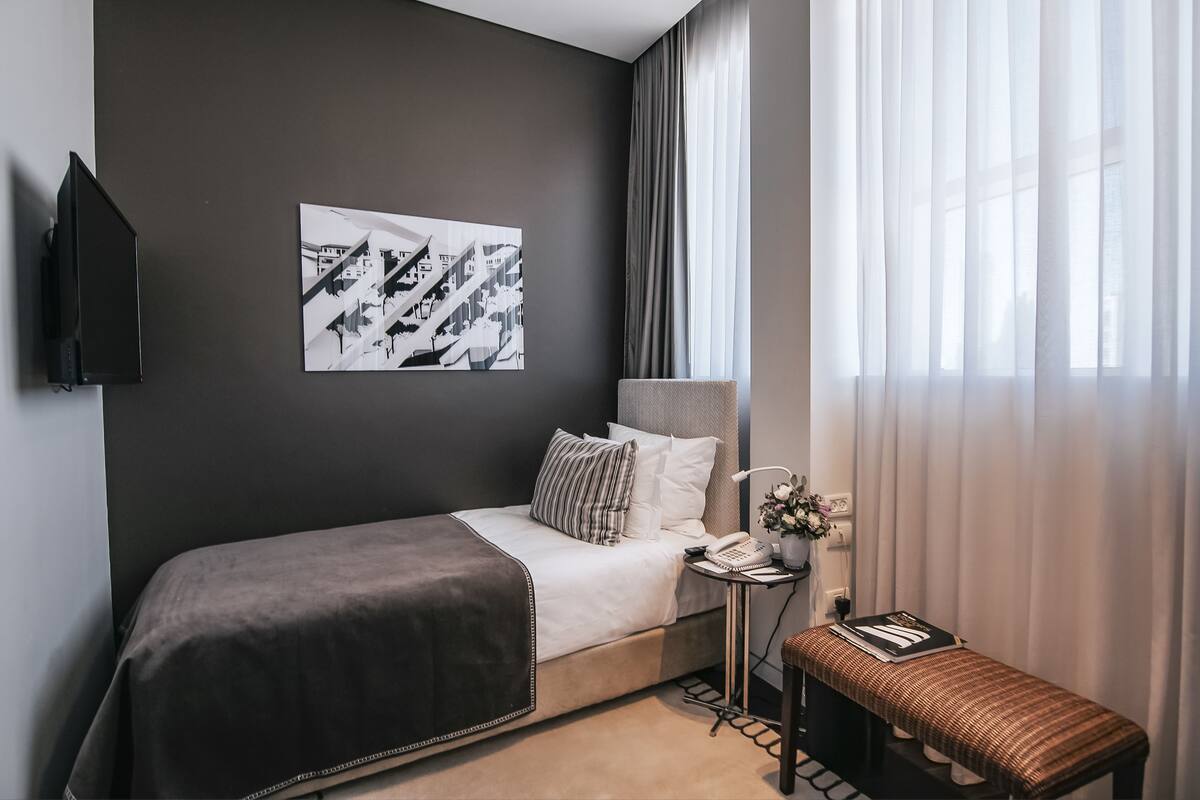 A hotel room with a bed, a bedside table with a lamp, a small stool, a wall-mounted TV, and a framed picture on the wall.