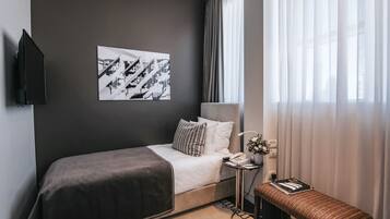 Una habitación de hotel con una cama, una mesita de noche con una lámpara, un taburete pequeño, un televisor montado en la pared y un cuadro enmarcado.