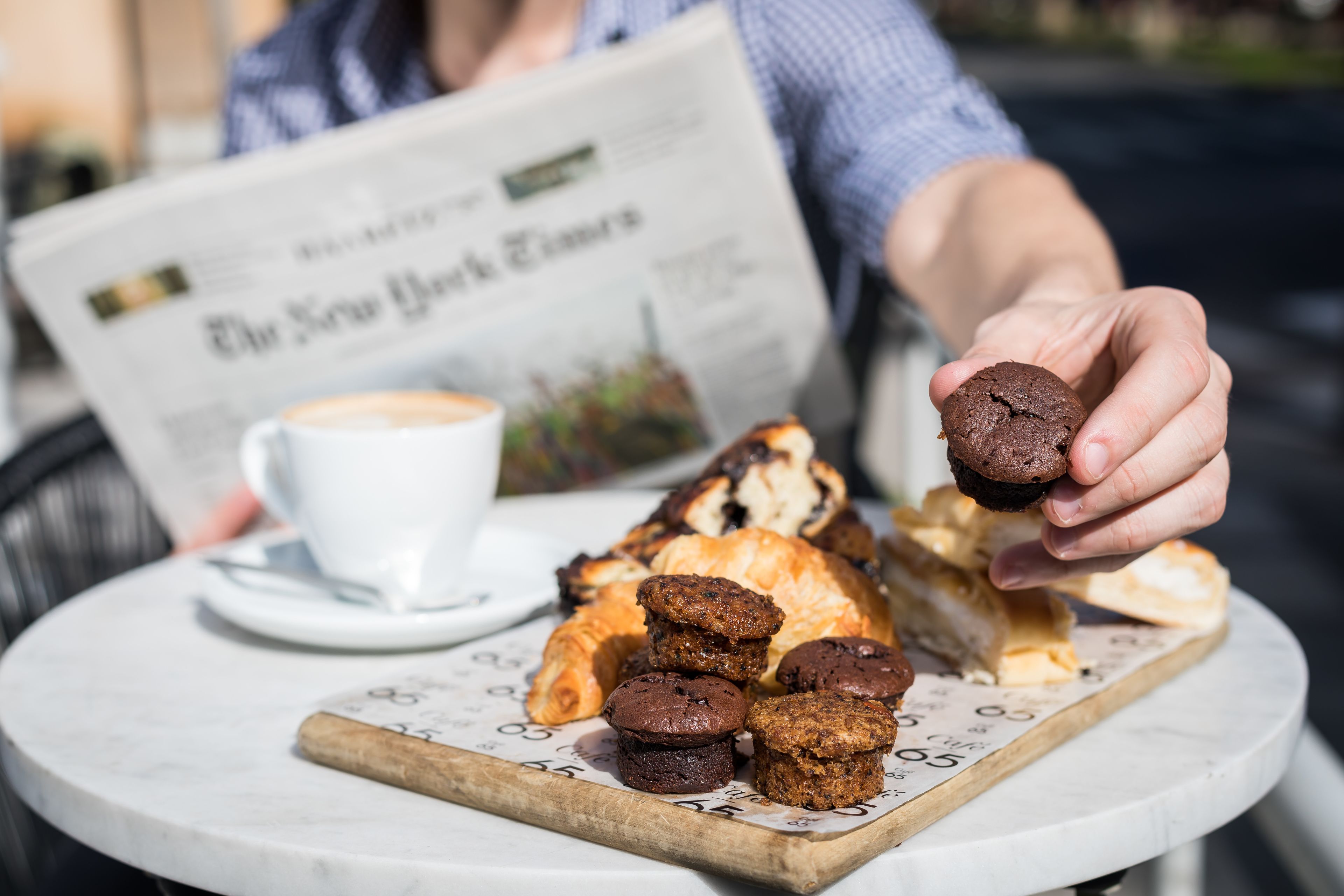 A table with pastries and a cup of coffee, a newspaper, and a person holding a muffin.