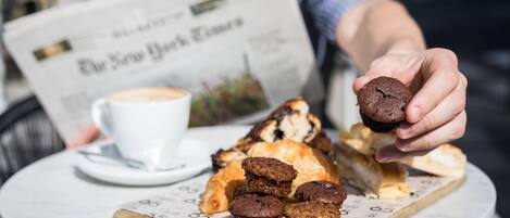 A table with pastries and a cup of coffee, a newspaper, and a person holding a muffin.