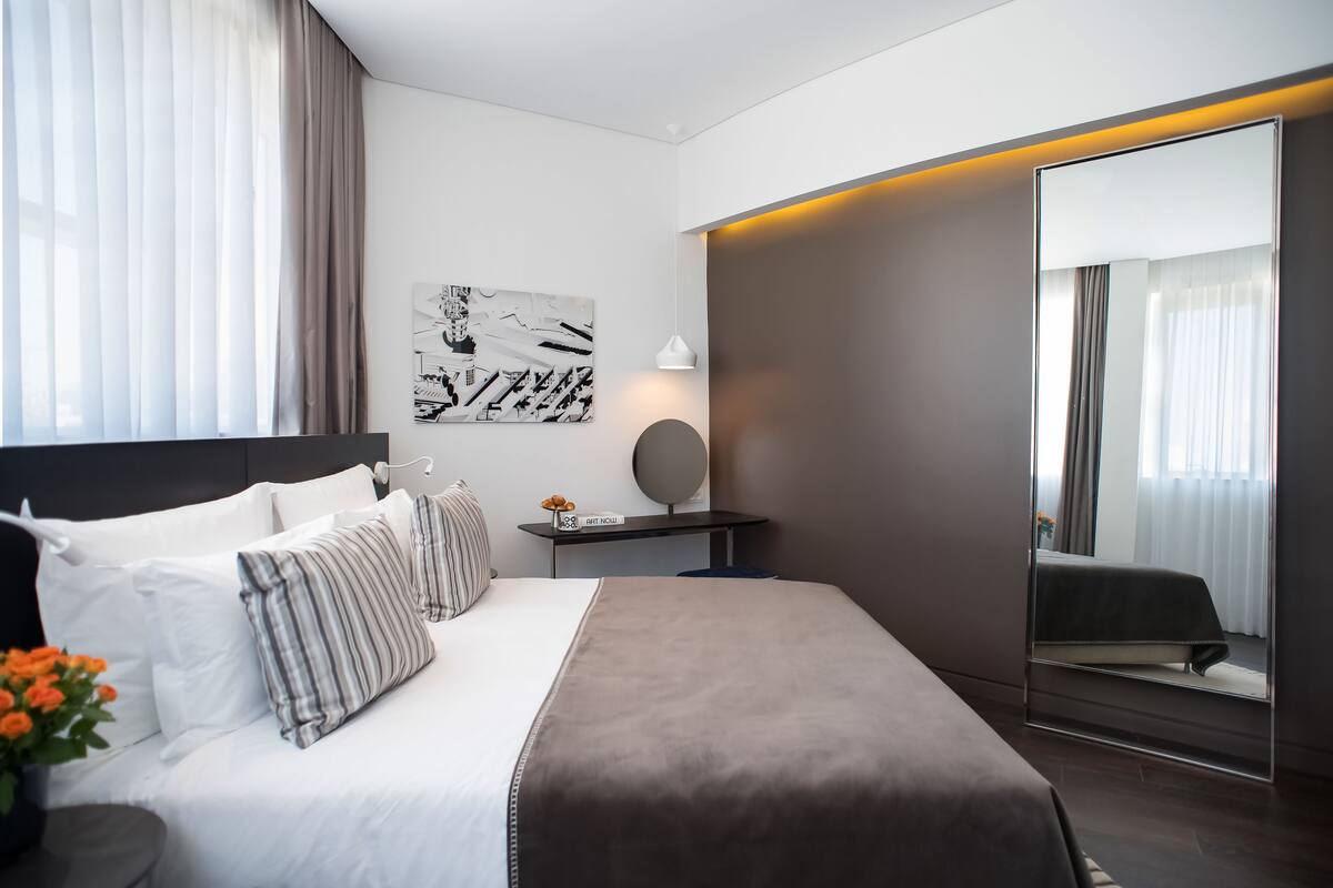 A modern hotel room with a large bed, a bedside table with a lamp, a mirror, and a painting on the wall.