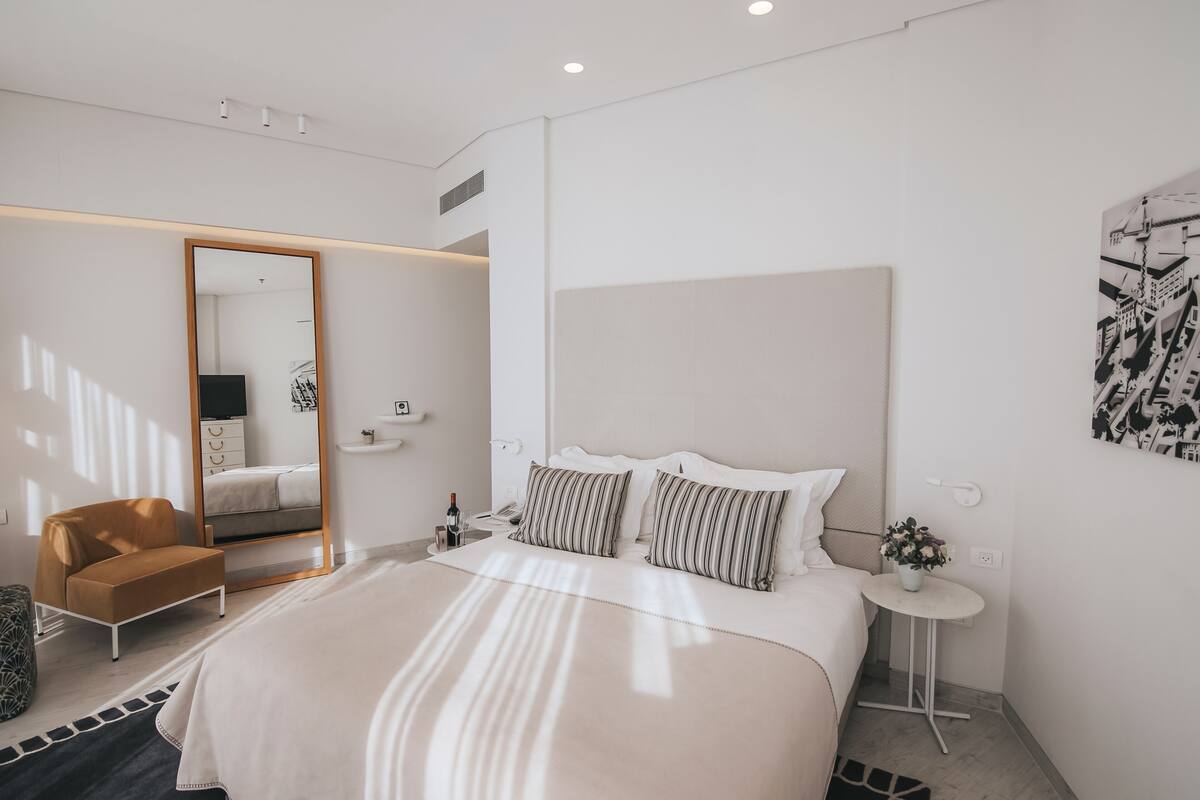 A modern hotel room with a large bed, a mirror, a small round table with a plant, and a painting on the wall.