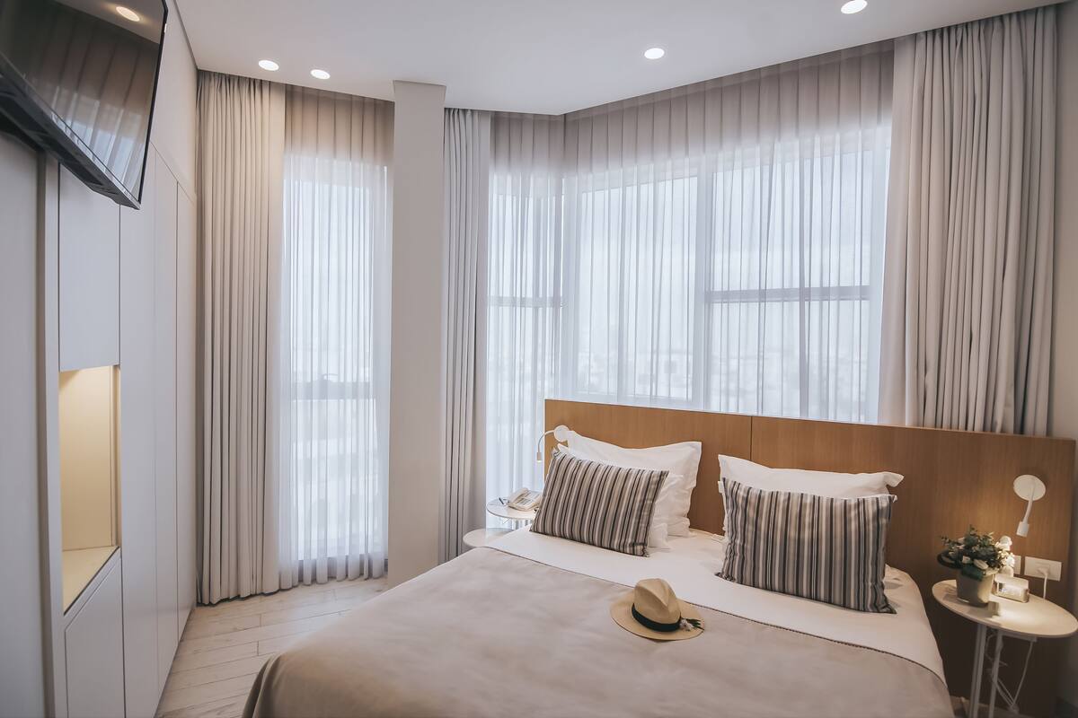 A modern hotel room with a large bed, two bedside tables, a TV mounted on the wall, and large windows with sheer curtains.