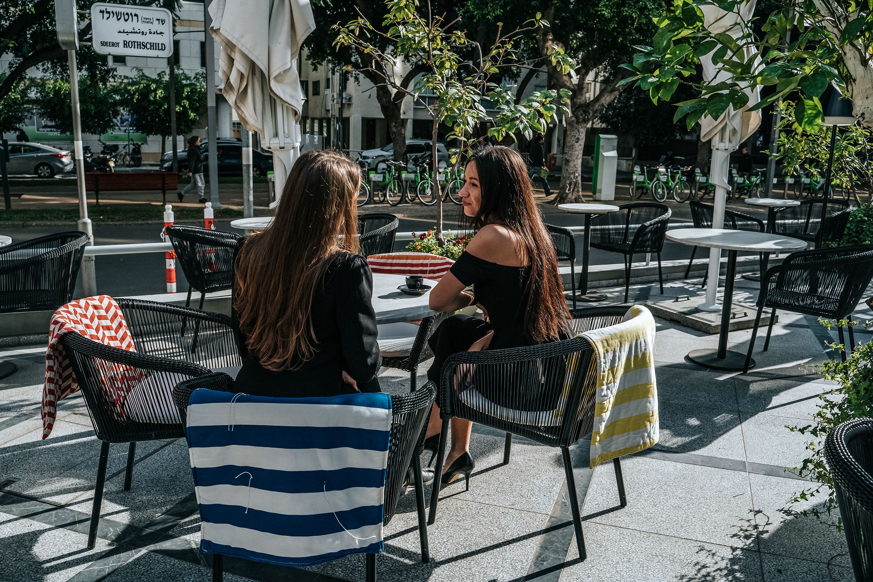 Dos mujeres sentadas en un café al aire libre con sillas y mesas de mimbre, rodeadas de árboles y vistas a la calle.
