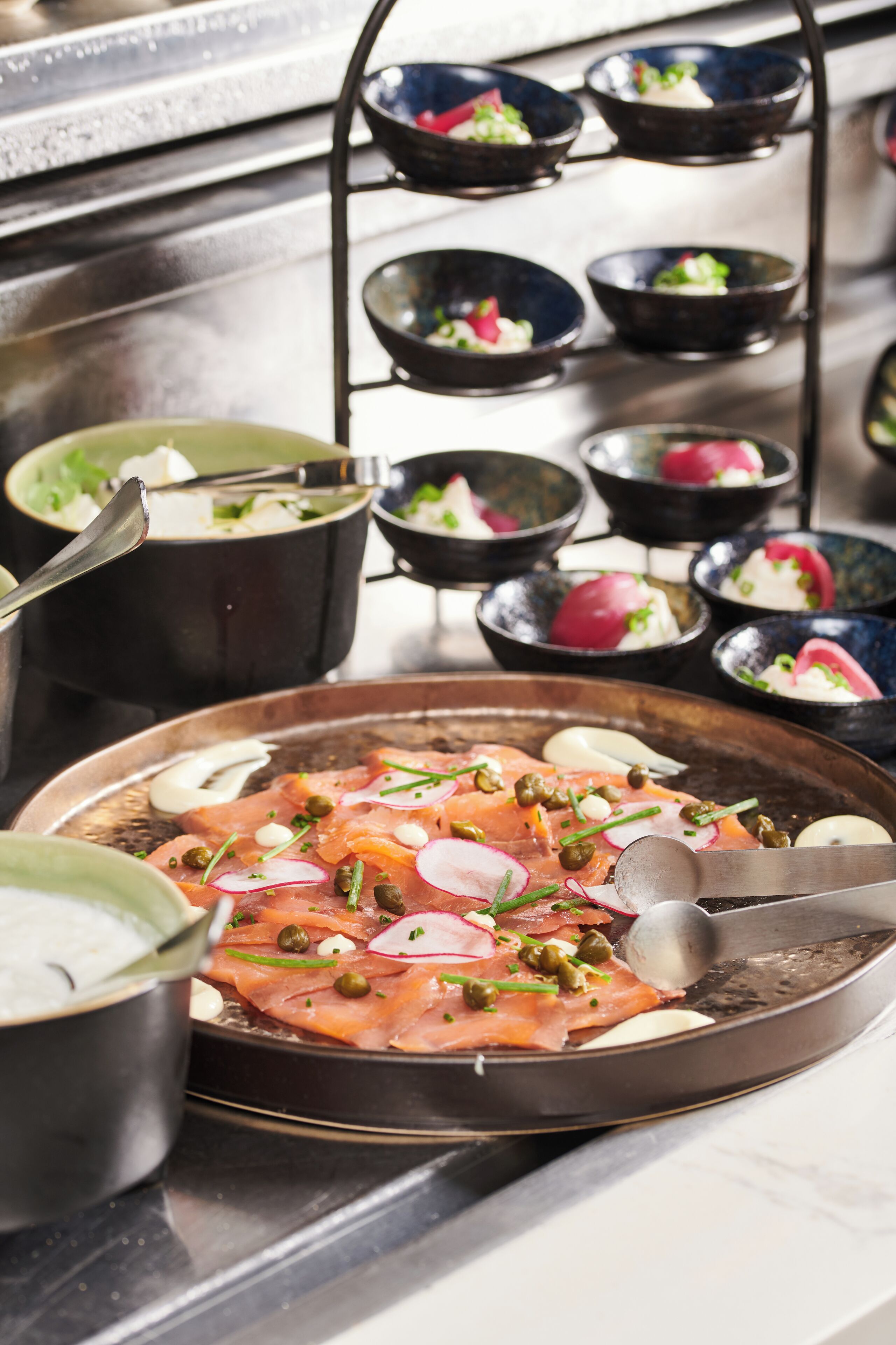 Uma mesa posta com diversos pratos, incluindo um prato de salmão defumado e alcaparras, e vários recipientes com diferentes alimentos.