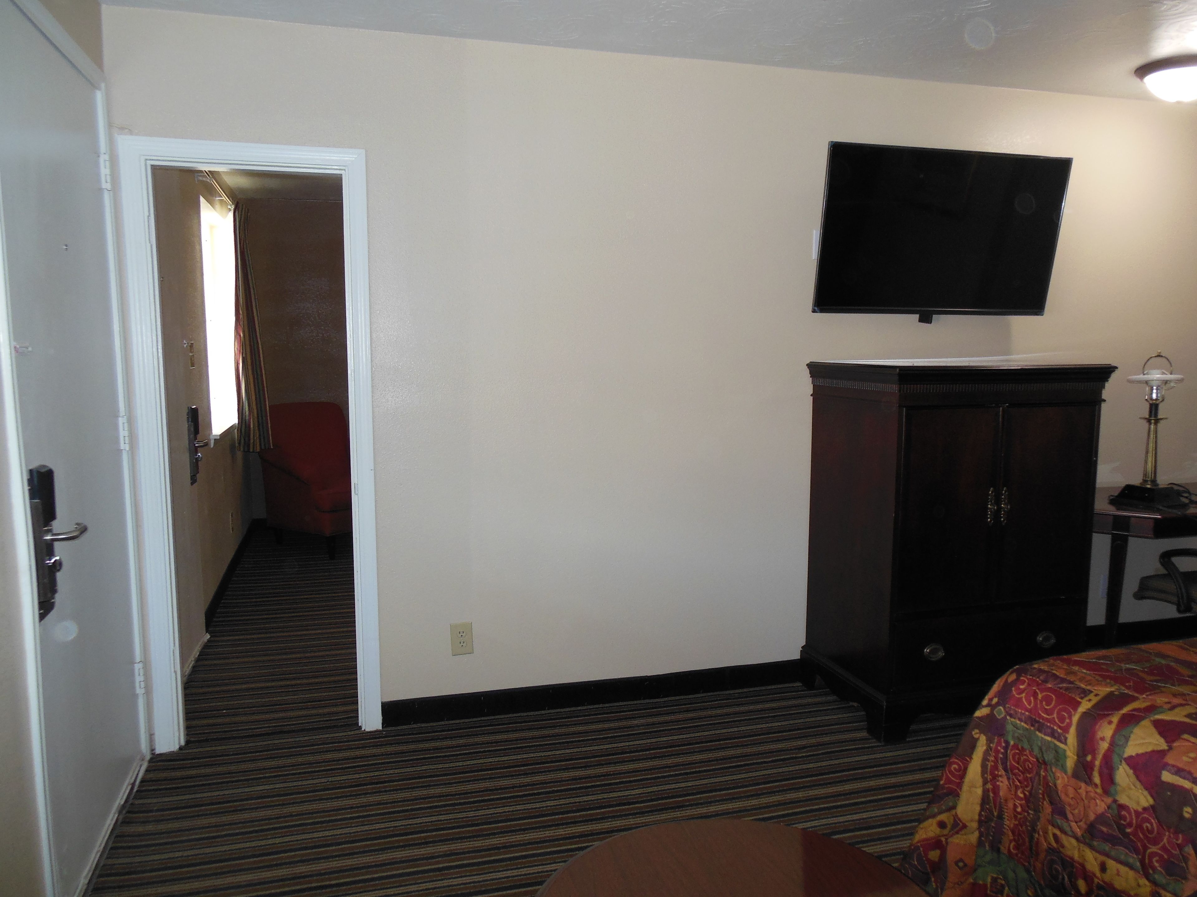Quarto de hotel com uma cama, televisão fixada na parede, um armário de madeira escura e piso coberto por carpete.