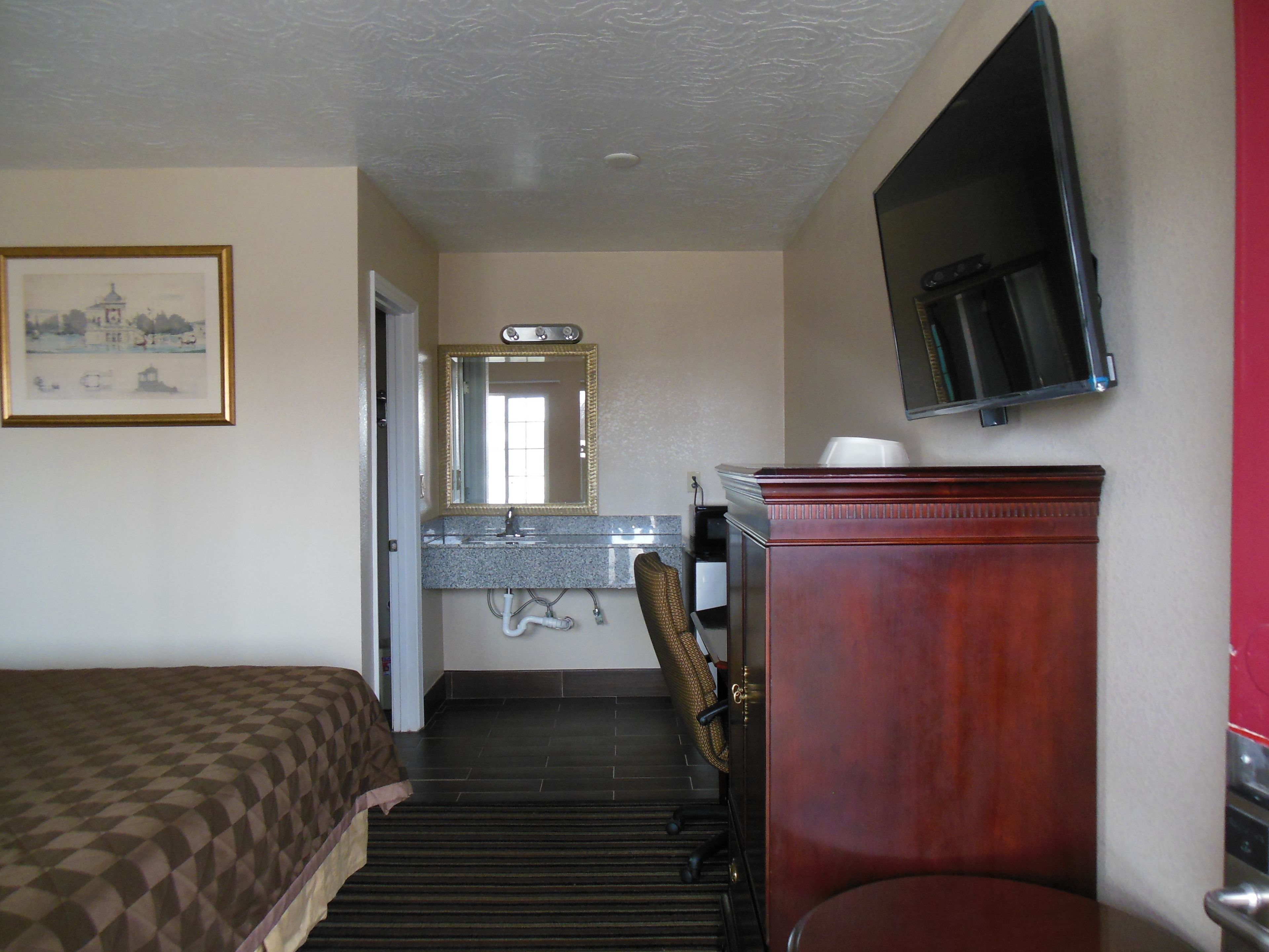 A hotel room with a bed, a bathroom with a sink and mirror, a television mounted on the wall, and a framed picture on the wall.