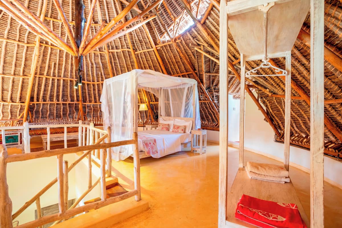 Quarto tradicional com telhado de colmo, uma cama, uma mesinha e uma cadeira suspensa.