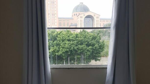 A hotel room with a bed, white curtains, and an air conditioning unit. - Hotel Pousada do Papa (Aparecida)