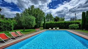 A rectangular swimming pool with clear blue water, surrounded by a gravel border. There are four lounge chairs with red cushions aligned along the left side of the pool. The pool is set in a landscaped area with various green shrubs and trees in the background. - Villa Fiamma (Capannori)