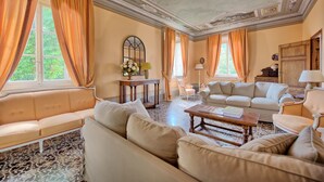 A spacious living room with a sofa, a coffee table, and a dining table. There are large windows with curtains, a chandelier, and a decorative ceiling. - Villa Fiamma (Capannori)