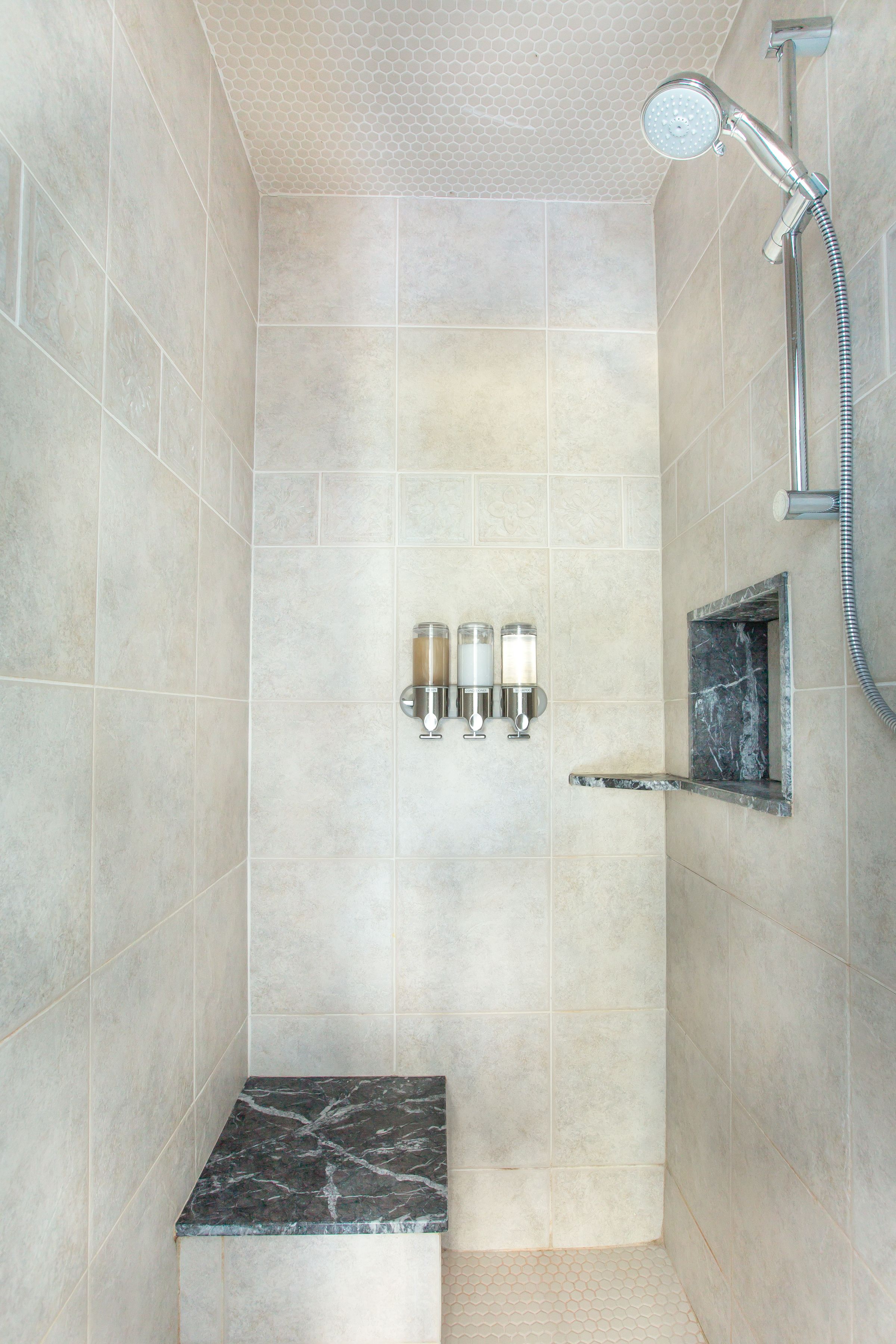 A shower area with a handheld showerhead, a wall-mounted soap dispenser, and a marble-topped sink.