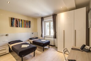 A hotel room with two single beds, a desk with a telephone, a chair, a window with curtains, and a colorful abstract painting on the wall. - The Bricks Rome (Rome)