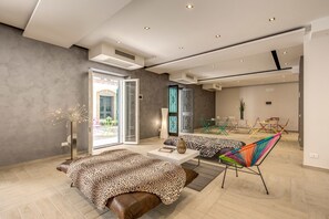 Una habitación de hotel moderna con una cama grande, un sofá y una silla colorida. Se ve un ventanal con vistas al exterior.