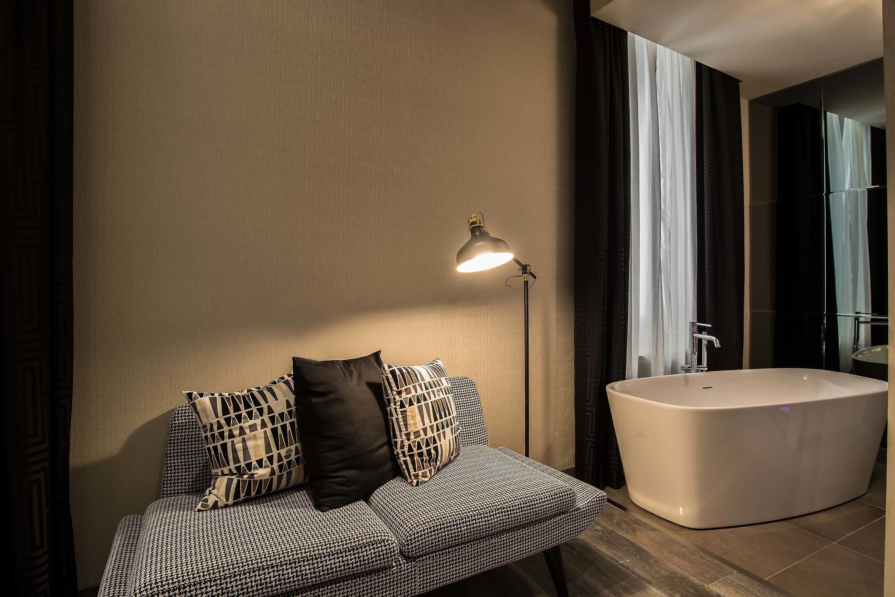 A modern bathroom with a freestanding bathtub, a sofa with patterned cushions, and a floor lamp.