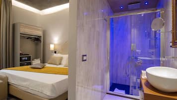 Una habitación de hotel moderna con una cama grande, un vestidor, un baño con cabina de ducha de vidrio y un lavamanos amplio.