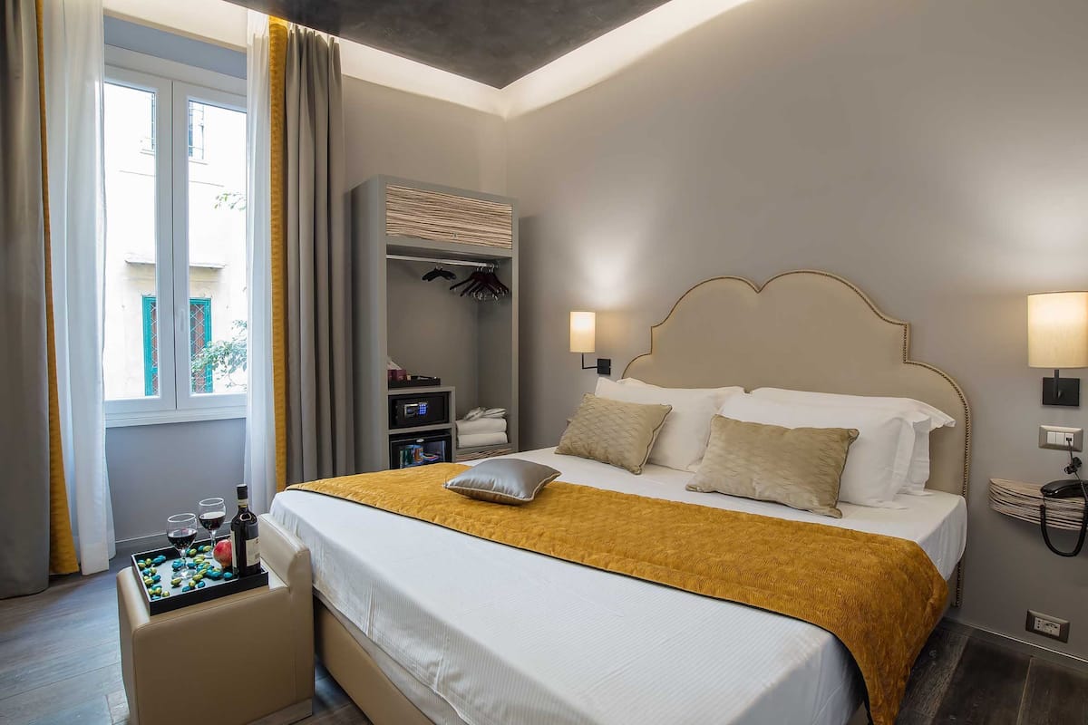 A hotel room with a bed, bedside lamps, a bedside table with a bottle and glasses, a wardrobe, and a window with curtains.