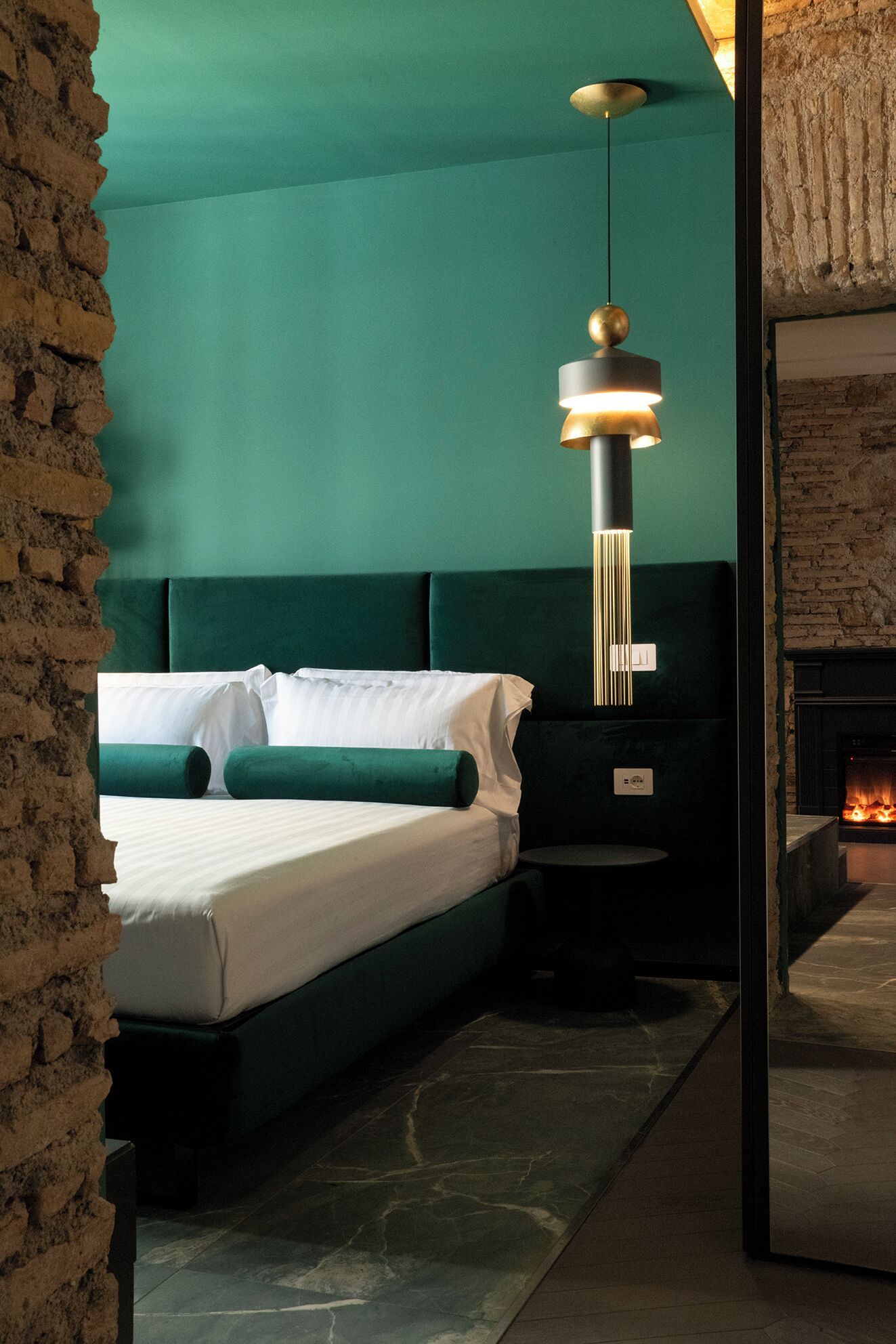 A modern bedroom with a green upholstered headboard, a bed with white linens, a hanging pendant light, and a visible fireplace.