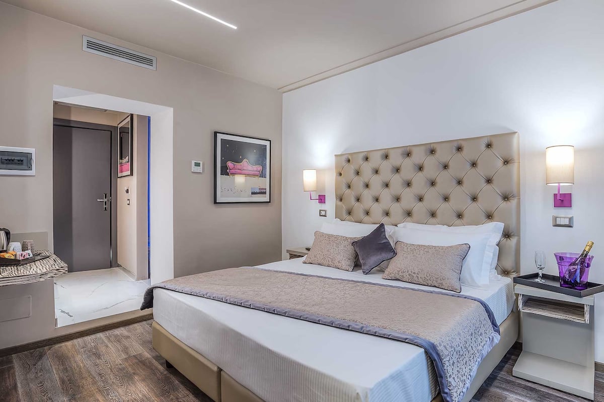 A modern hotel room with a large bed, a bedside table with a lamp, a small side table with a drink, and a framed picture on the wall.