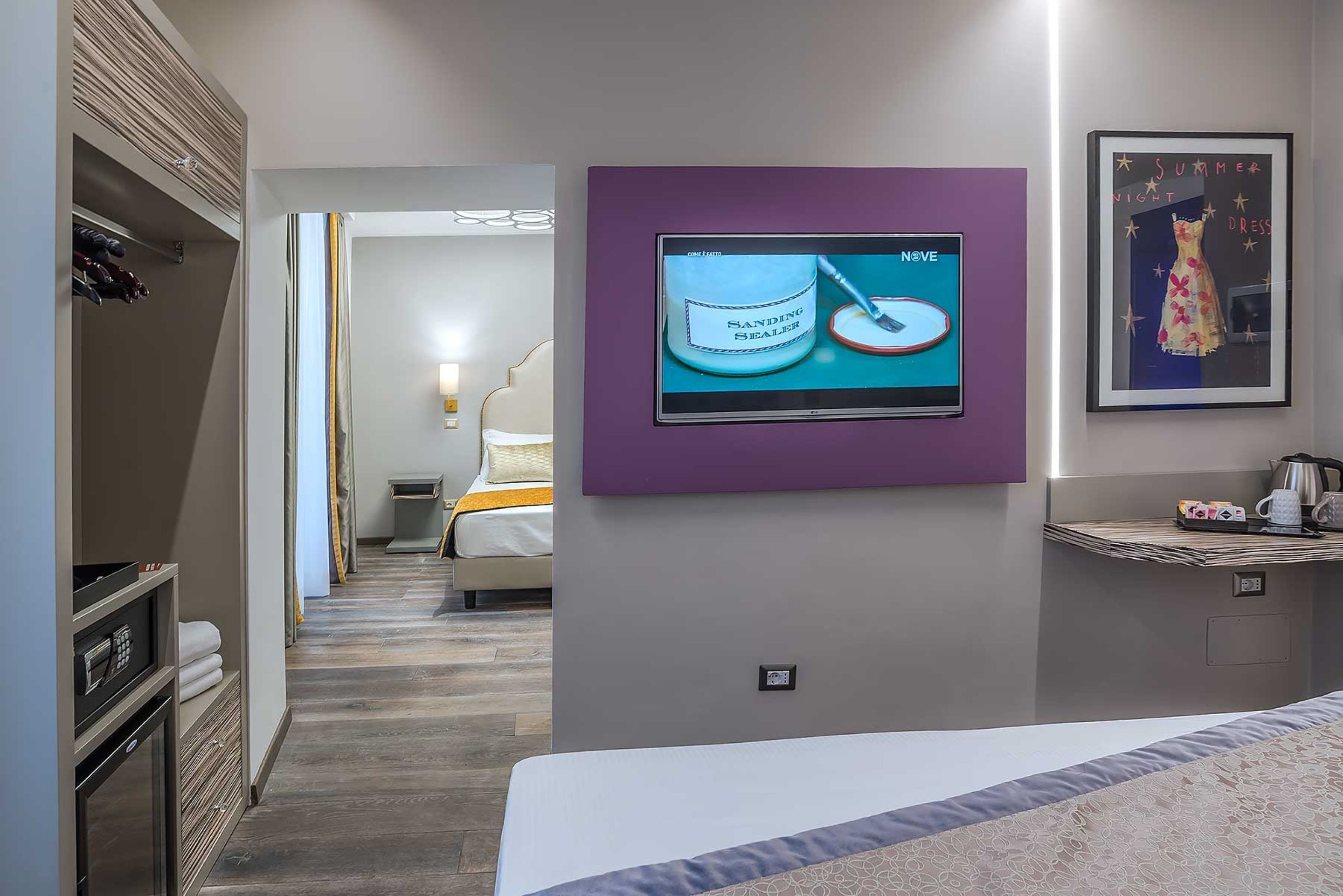 A hotel room with a bed, a television mounted on the wall, a framed picture, and a small shelf with items.