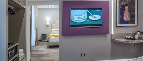 A hotel room with a bed, a television mounted on the wall, a framed picture, and a small shelf with items.