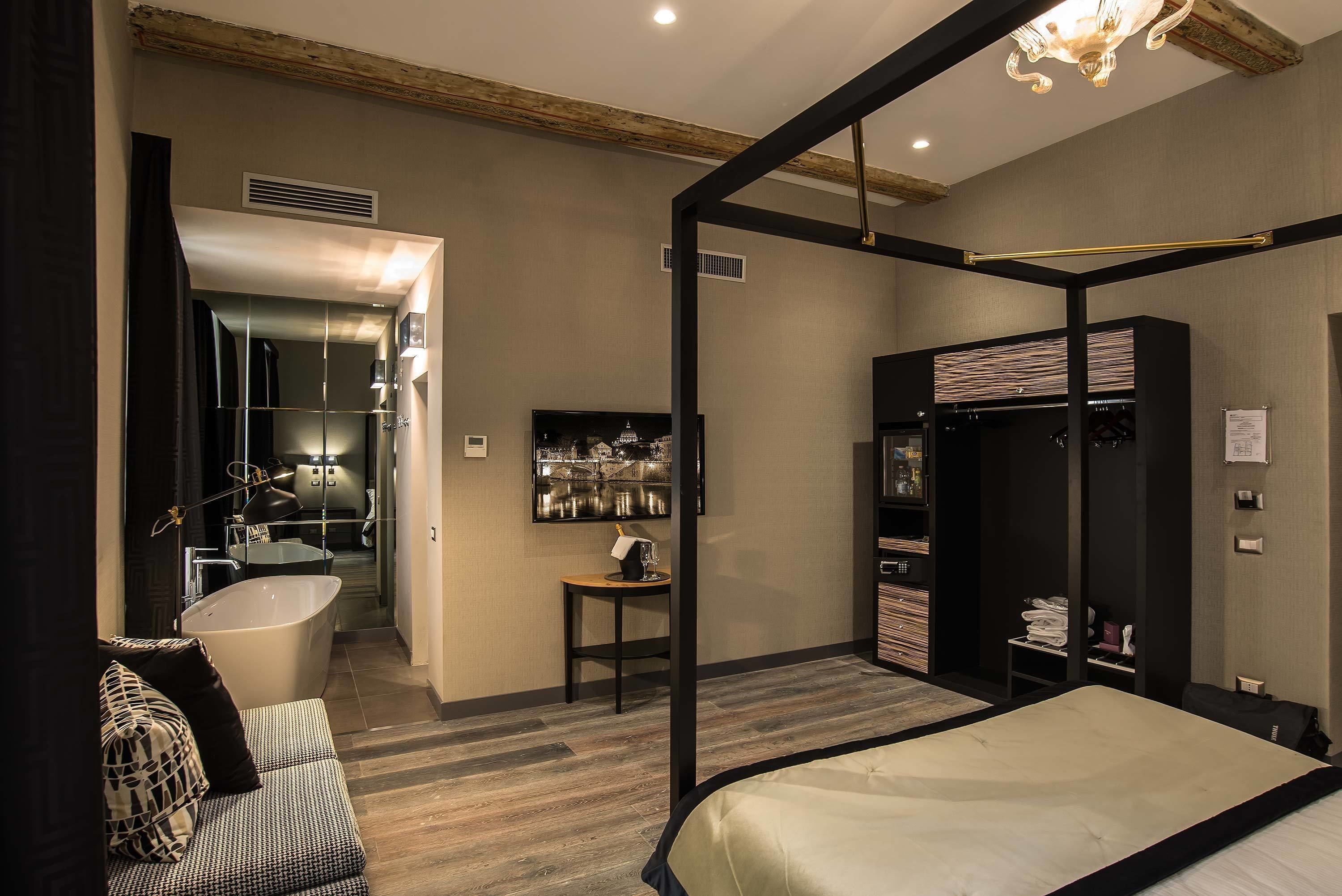 A modern hotel room with a large bed, a separate bathroom with a bathtub, a wardrobe, and a small table with a lamp.