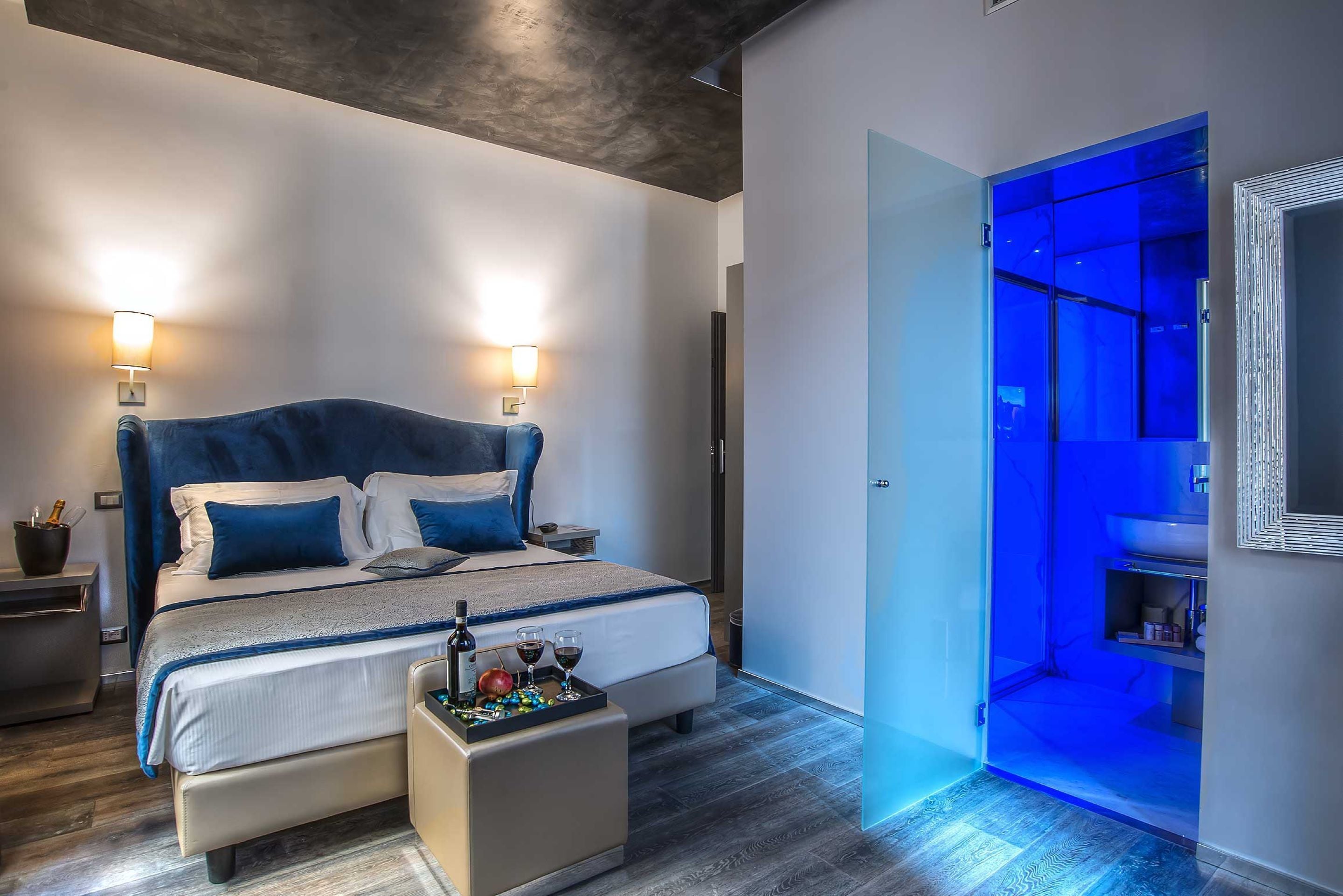A modern bedroom with a large bed, bedside lamps, a small table with drinks, and a blue-lit bathroom.