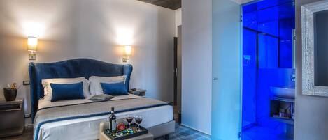 A modern bedroom with a large bed, bedside lamps, a small table with drinks, and a blue-lit bathroom.