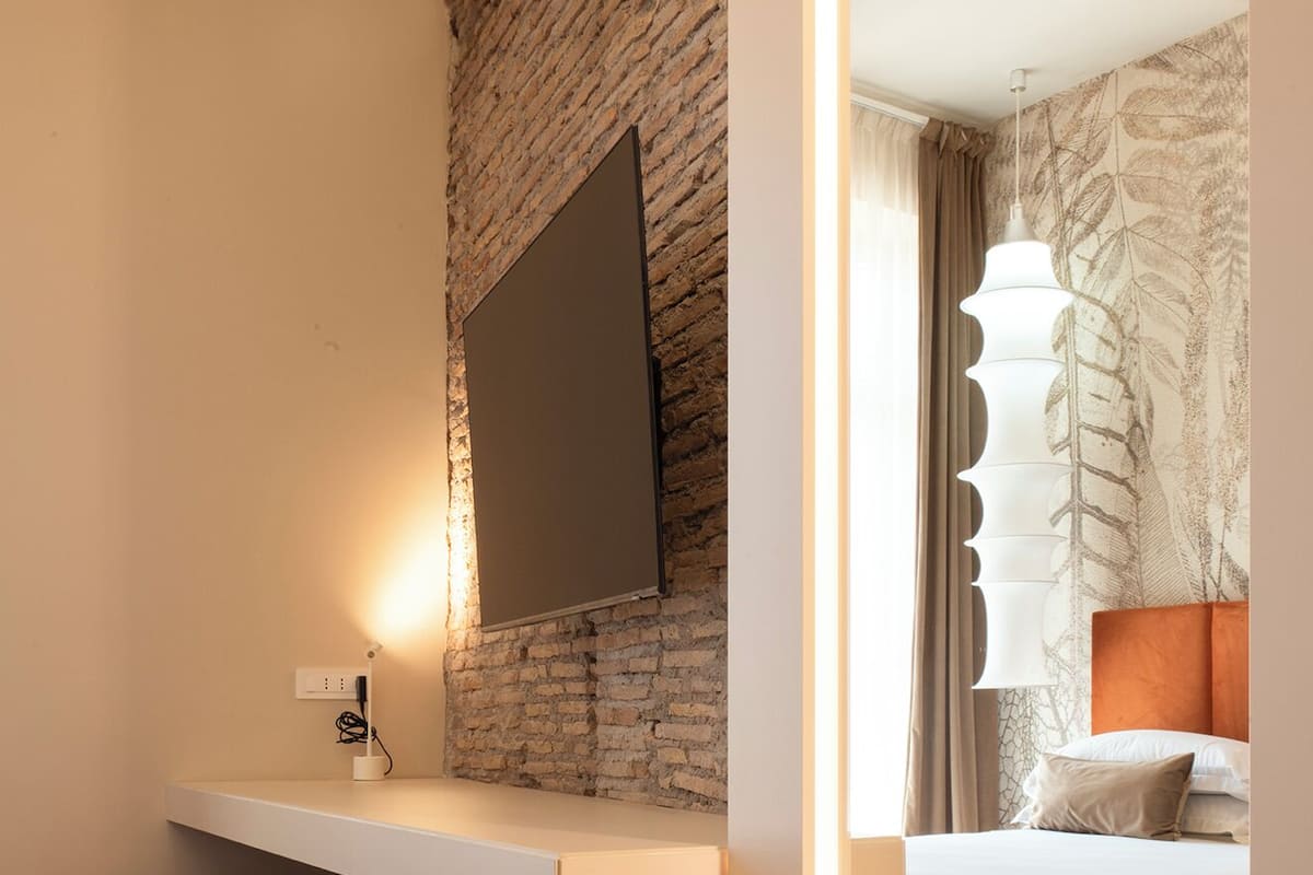 A modern hotel room with a brick accent wall, a flat-screen TV mounted above a white desk, a red chair, and a bed with a patterned headboard.