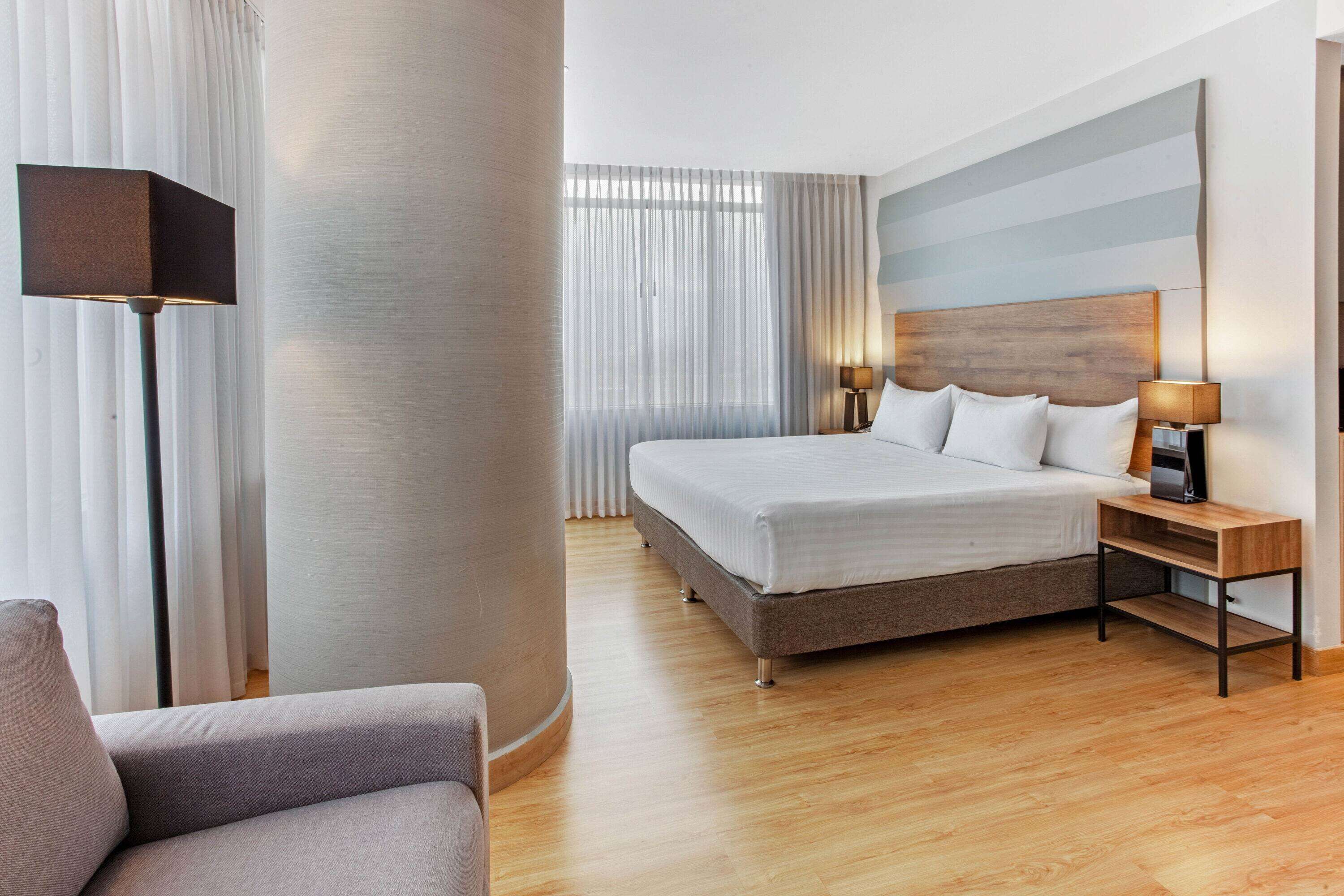 Een moderne hotelkamer met een groot bed, een nachtkastje met een lamp en uitzicht op de stad door dunne gordijnen.