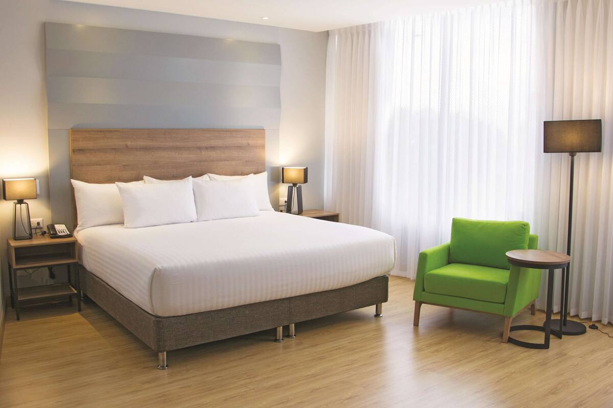 A hotel room with a large bed, two bedside tables with lamps, a green armchair, and a floor lamp.