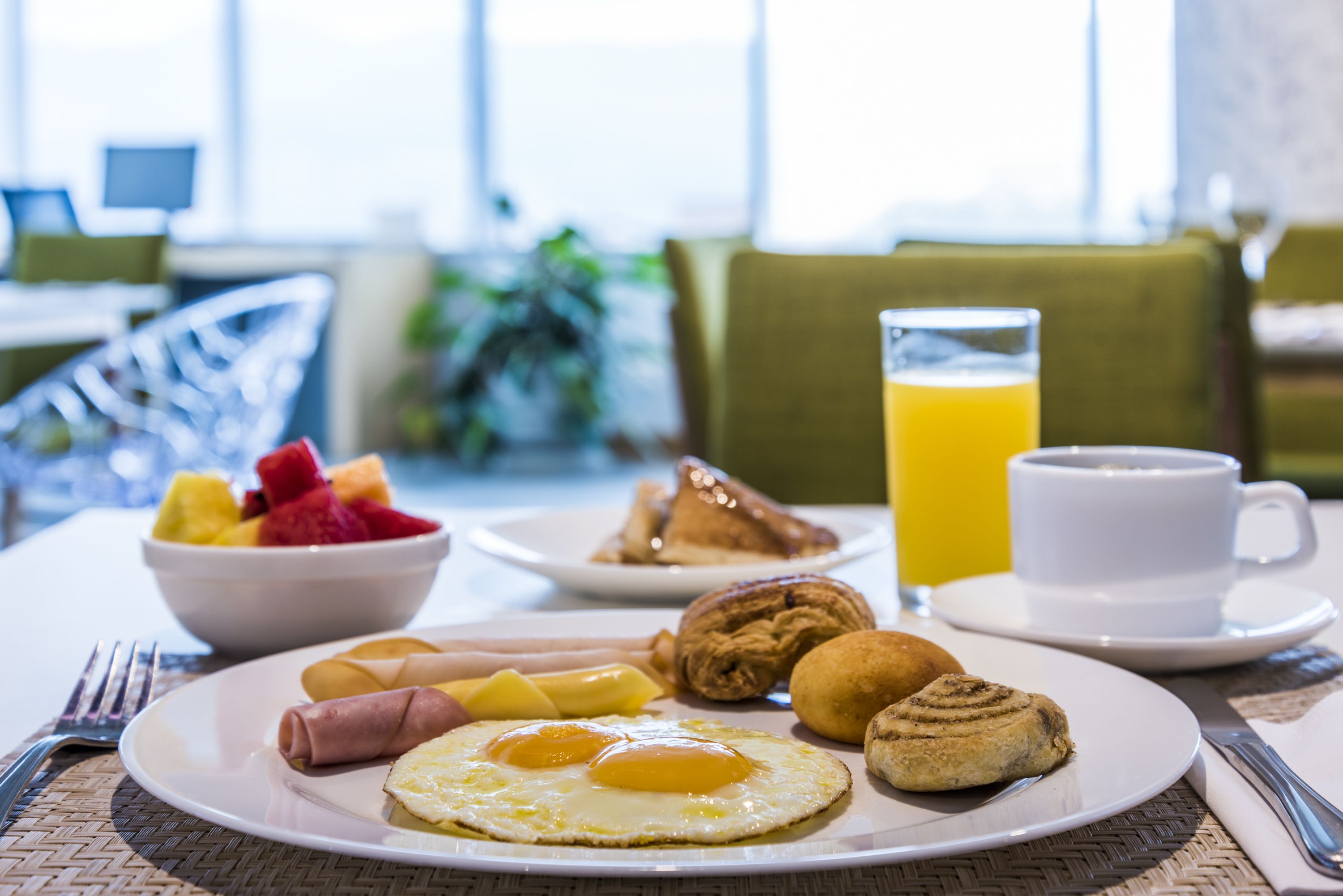Un desayuno con un plato de huevos, bollería y fruta, un vaso de zumo de naranja y una taza de café sobre una mesa.