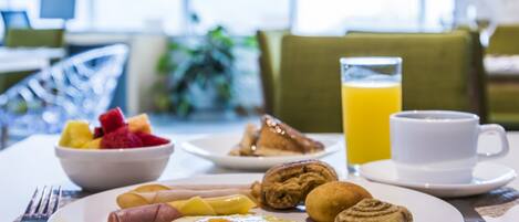 Un desayuno con un plato de huevos, bollería y fruta, un vaso de zumo de naranja y una taza de café sobre una mesa.