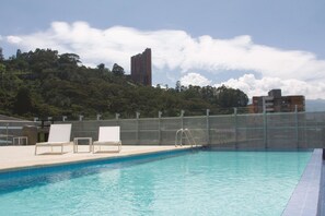 Una piscina en la azotea con sillones de descanso, una valla de seguridad y vistas a una colina boscosa y un edificio alto al fondo.