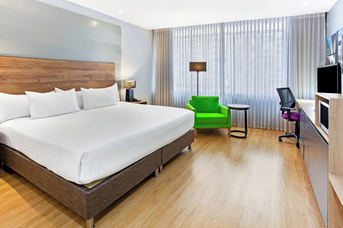 A hotel room with a large bed, a green armchair, a desk with a computer, and a TV.