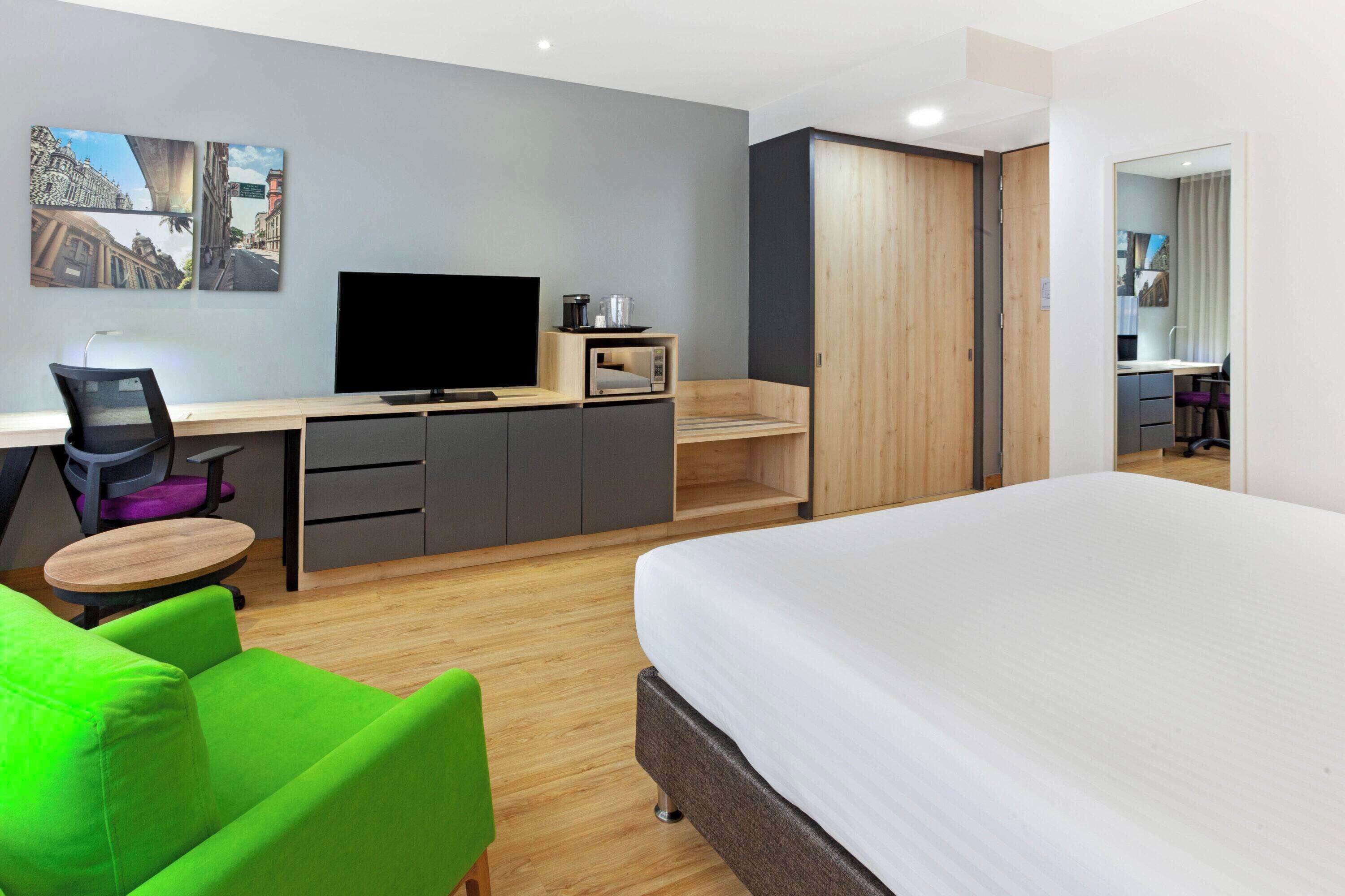Una habitación de hotel moderna con una cama, un escritorio con una computadora, una silla, un televisor y un armario.