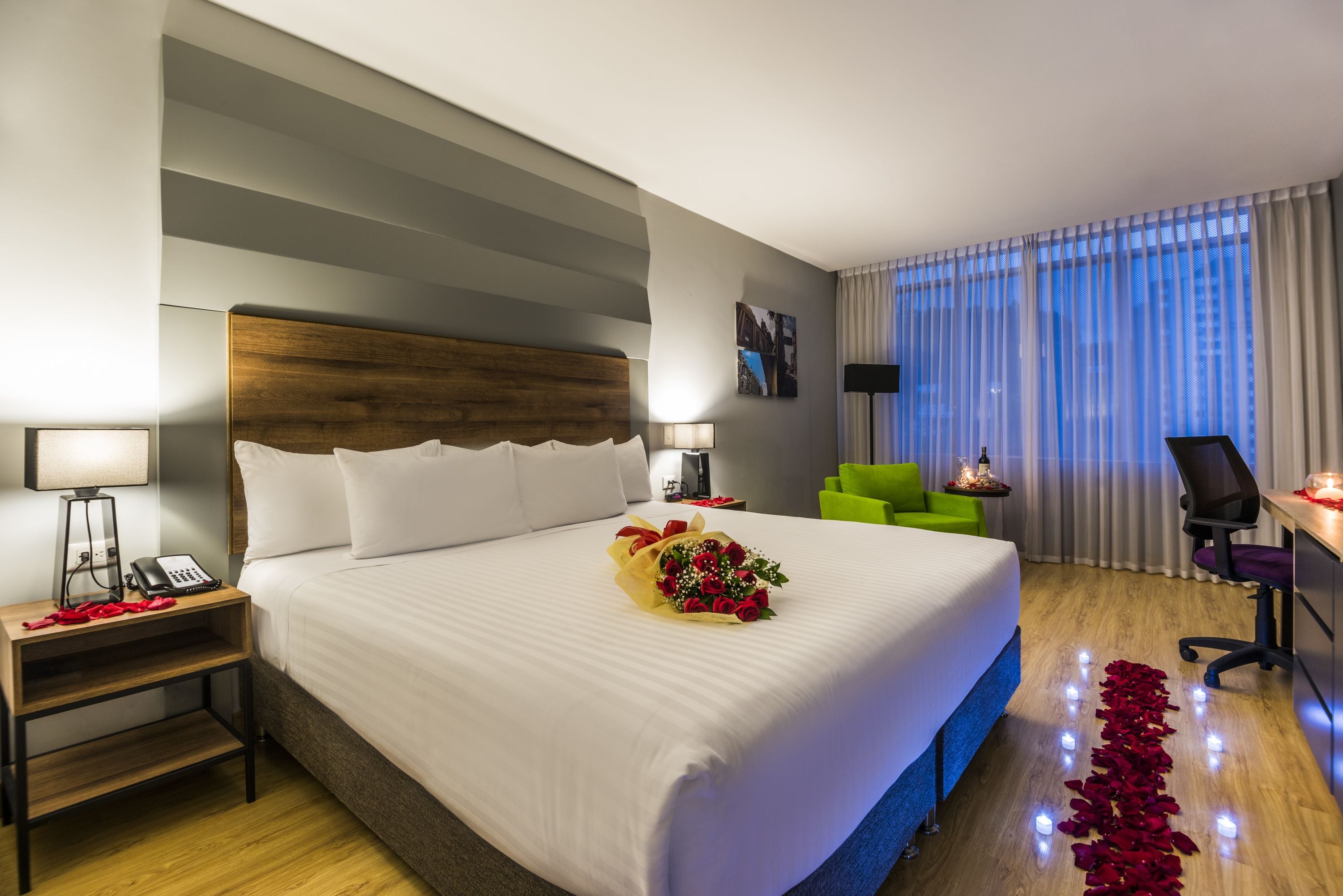 A hotel room with a large bed, bedside tables with lamps, a desk with a chair, and a view of the city through the window.