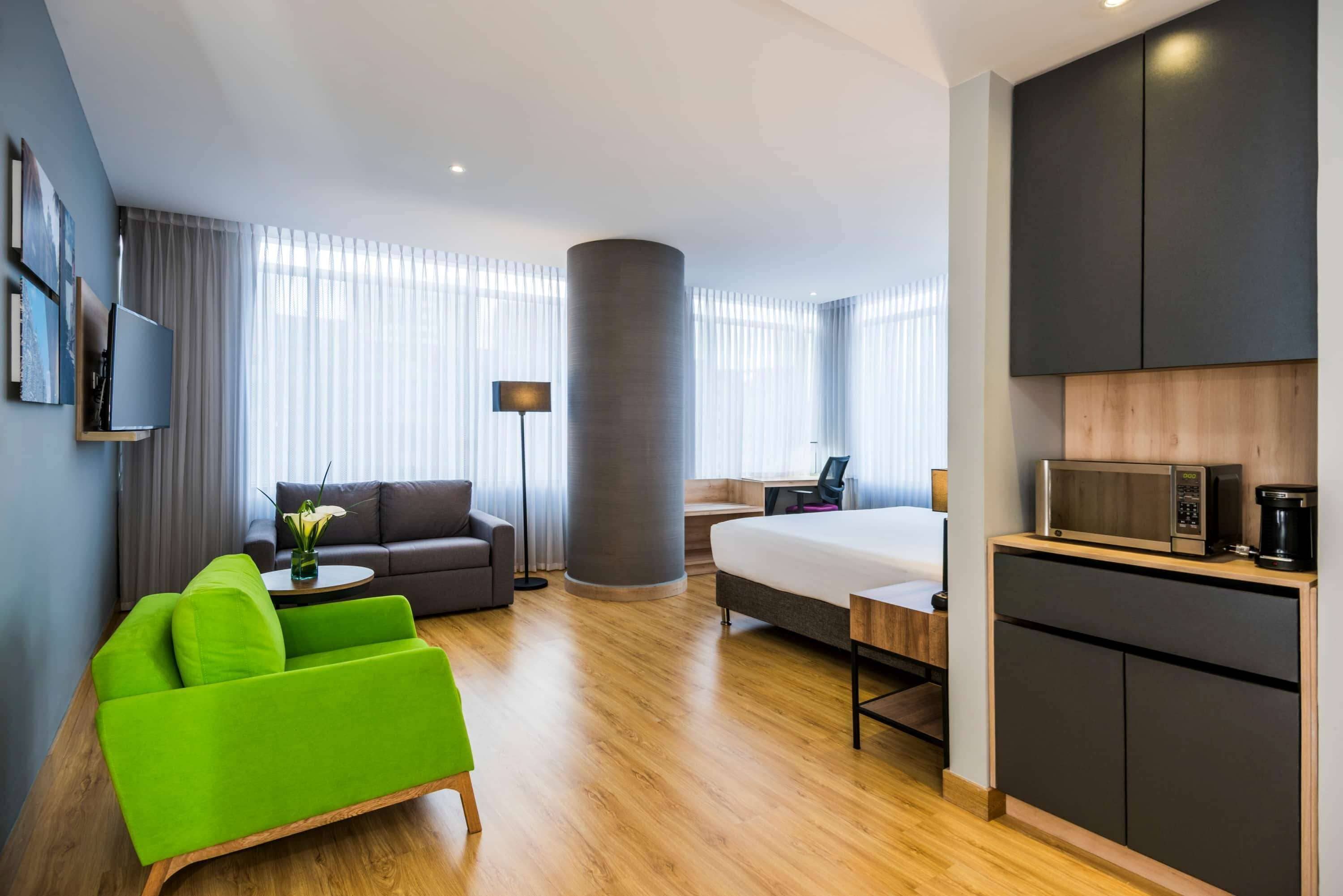 Una habitación de hotel moderna con piso de madera, un sillón verde, un sofá, una cama, un escritorio, un televisor de pantalla plana, una lámpara y un microondas.