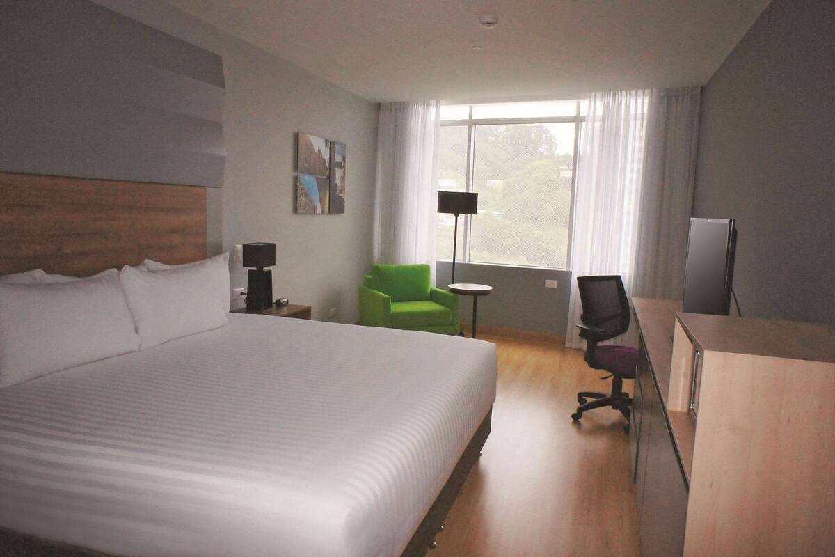 A hotel room with a large bed, a desk, a green chair, a lamp, and a window with curtains.