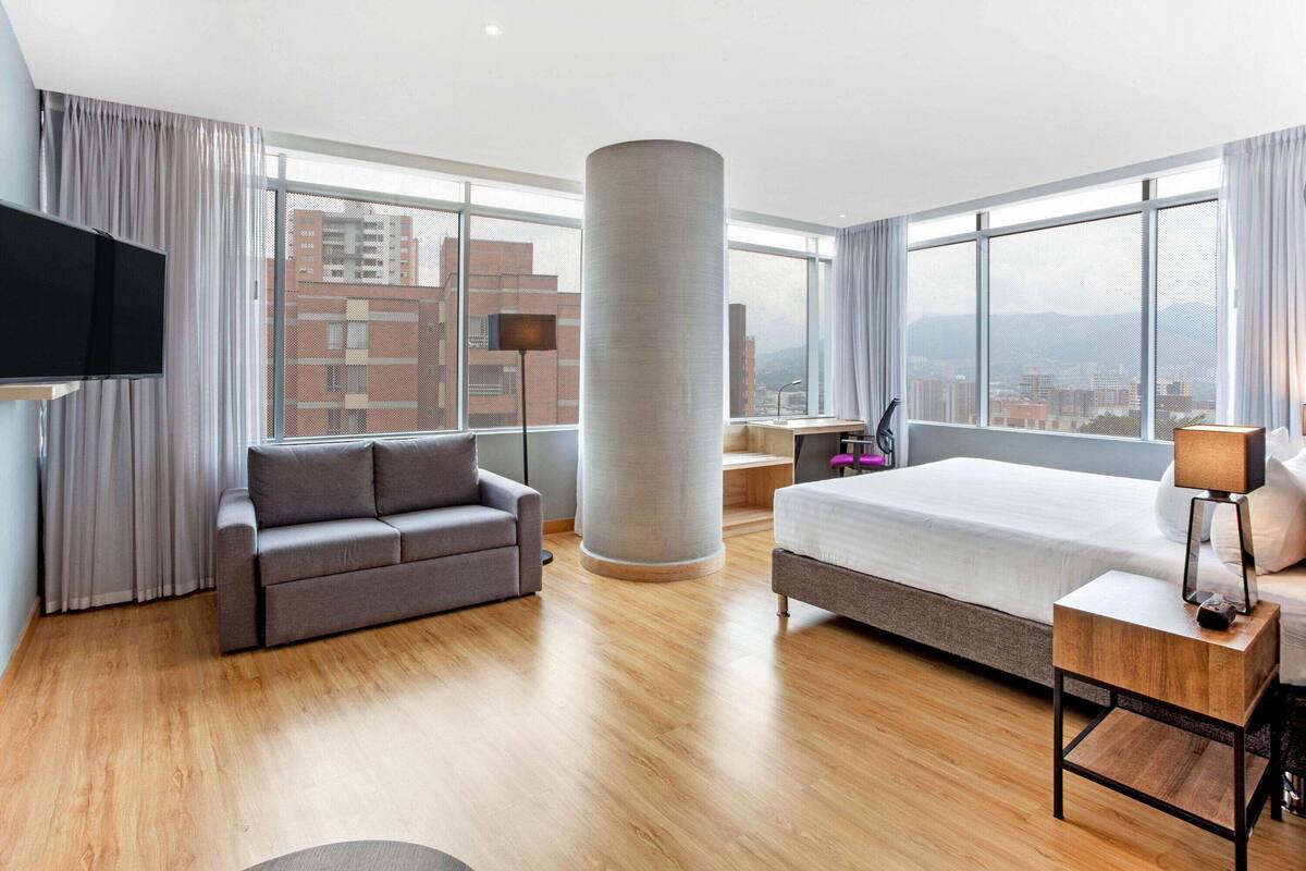 A modern hotel room with a large bed, a grey sofa, a wooden floor, a large window with a city view, and a flat-screen TV.