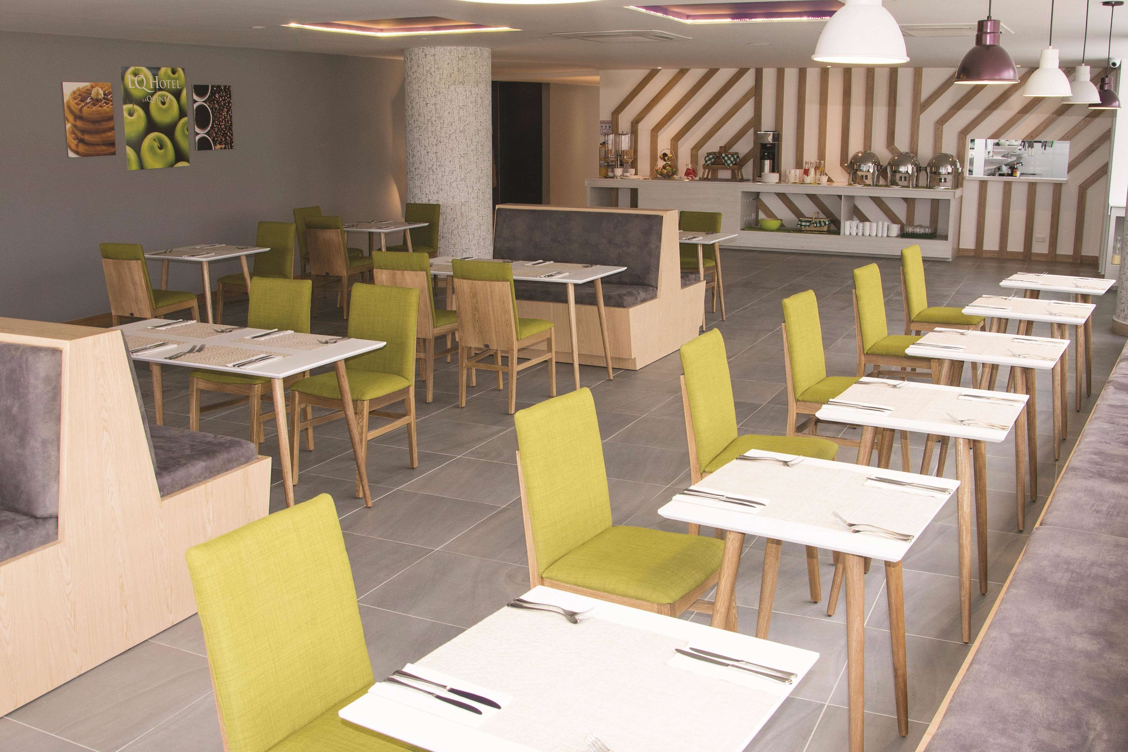 Een modern restaurant met groene en houten stoelen, witte tafels en een wand met strepen.