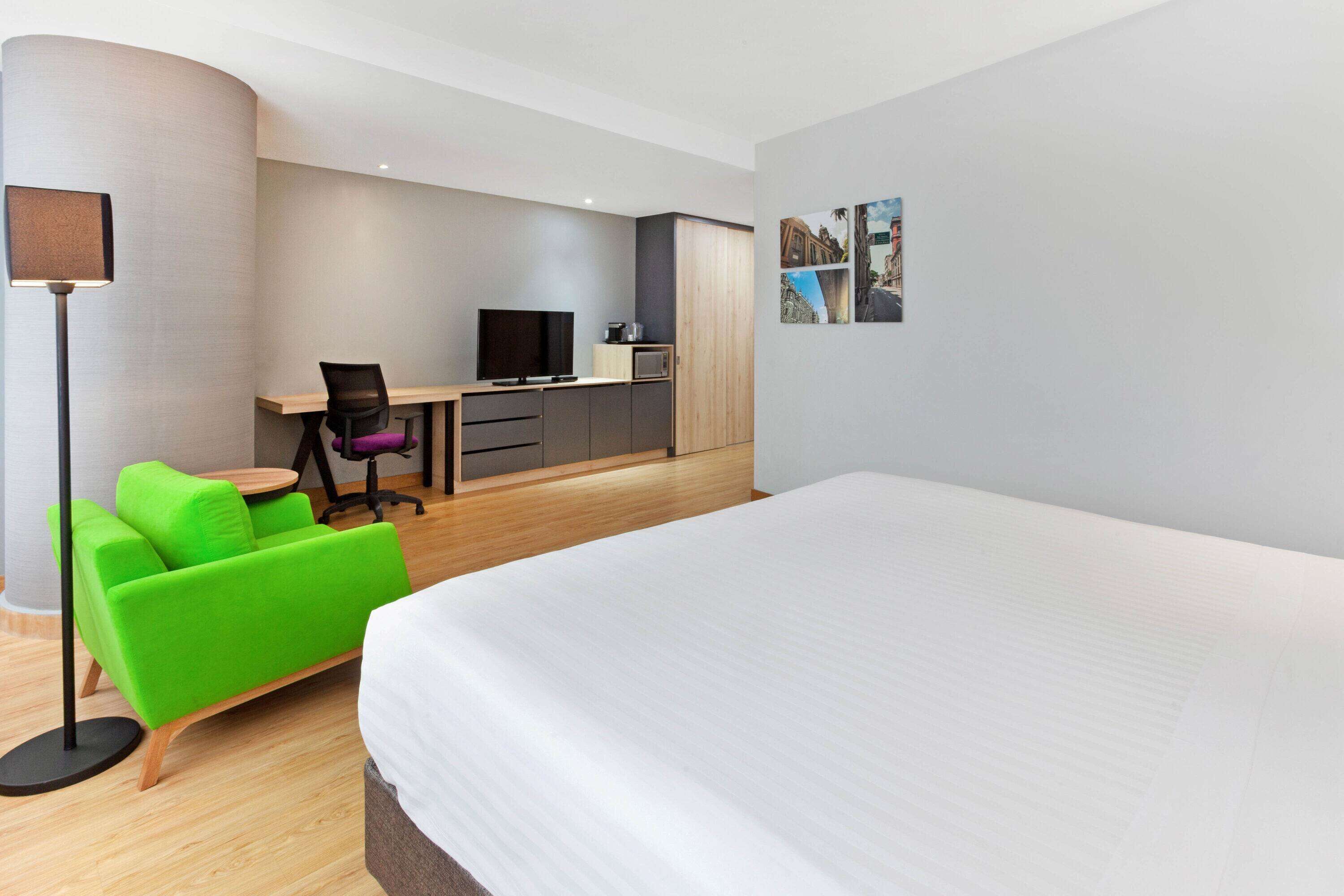 Una habitación de hotel moderna con una cama grande, un sofá verde, un escritorio con computadora y un televisor de pantalla plana.