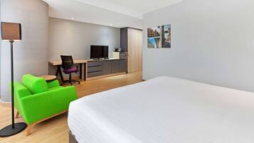 A modern hotel room with a large bed, a green sofa, a desk with a computer, and a flat-screen TV.