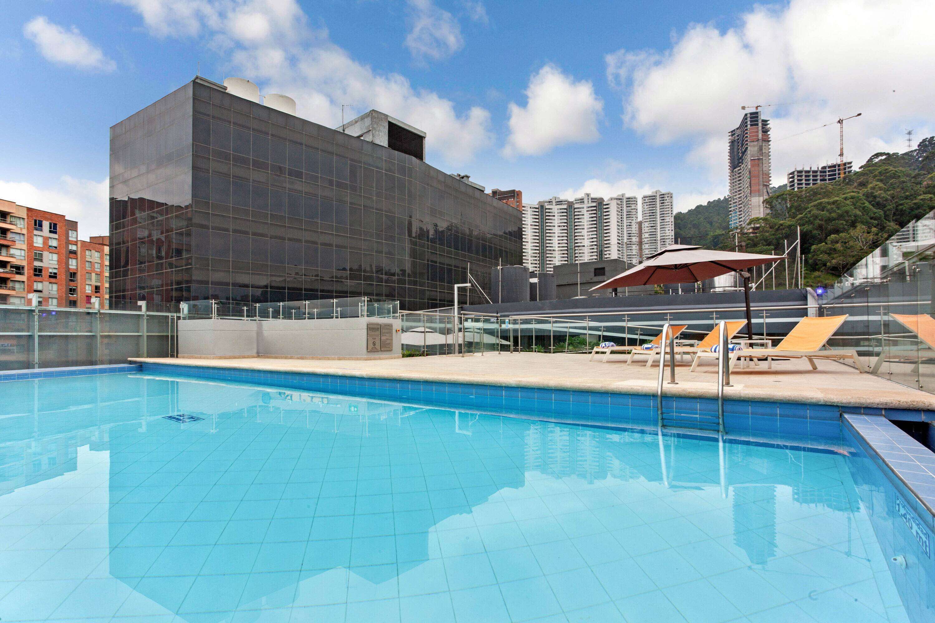 A swimming pool with lounge chairs and umbrellas, surrounded by modern buildings and a clear sky.