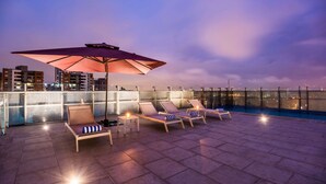 A rooftop terrace with lounge chairs and a large umbrella, overlooking a cityscape at dusk. - La Quinta by Wyndham Medellin (Medellín)