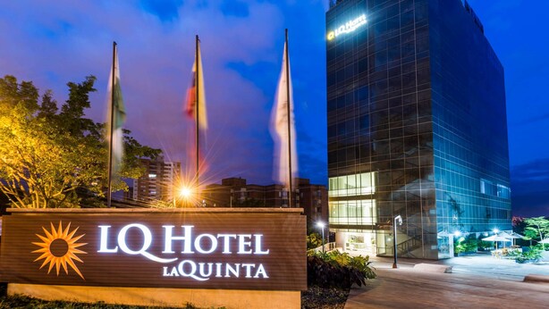 A hotel sign with a sun emblem, a modern glass building, and flags. - La Quinta by Wyndham Medellin (Medellín)